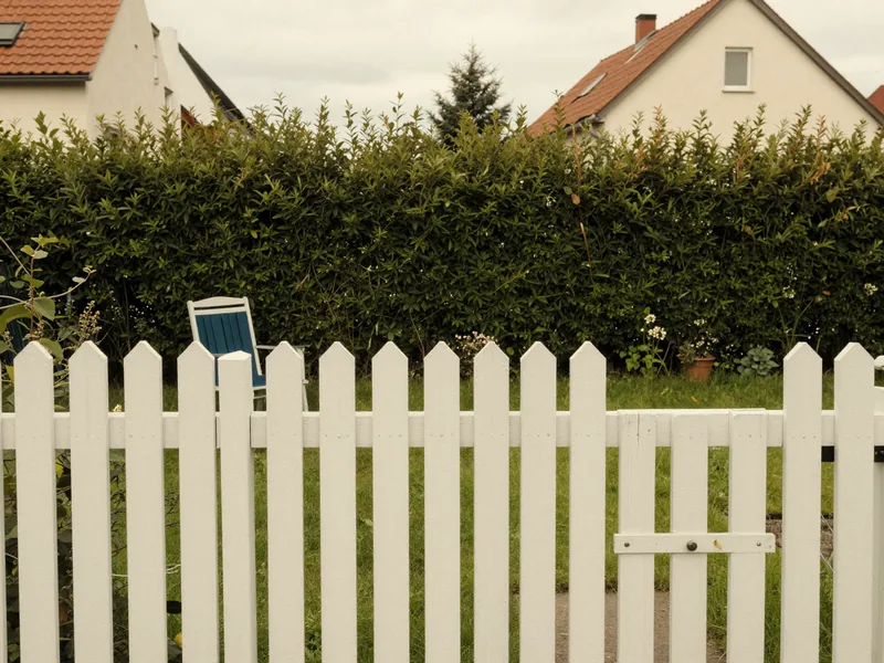 Blick ueber einen Gartenzaun auf eine hohe Hecke zwischen zwei Grundstuecken.